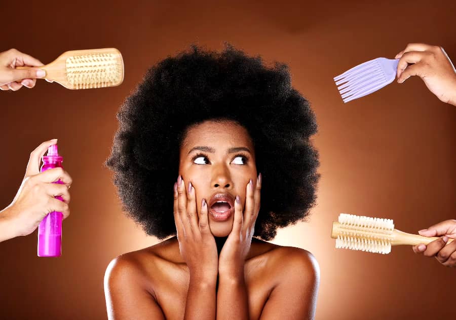A woman with a large afro looks surprised as hands around her hold hairbrushes, a wide-tooth comb, and a spray bottle against a brown background.